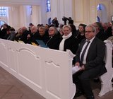 20 gut gekleitete Personen sitzen in mehreren Kirchenbänken hintereinander.