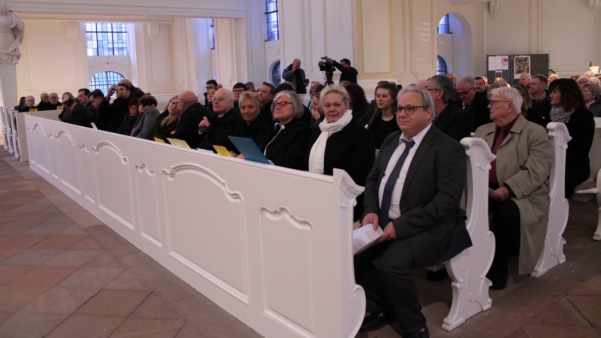 20 gut gekleitete Personen sitzen in mehreren Kirchenbänken hintereinander.