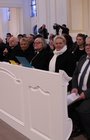 20 gut gekleitete Personen sitzen in mehreren Kirchenbänken hintereinander.